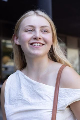 Smiling young woman glancing right in white one-shoulder top with brown leather strap