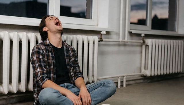Yawning young man sitting on floor against radiator in moody urban studio at dusk solitary