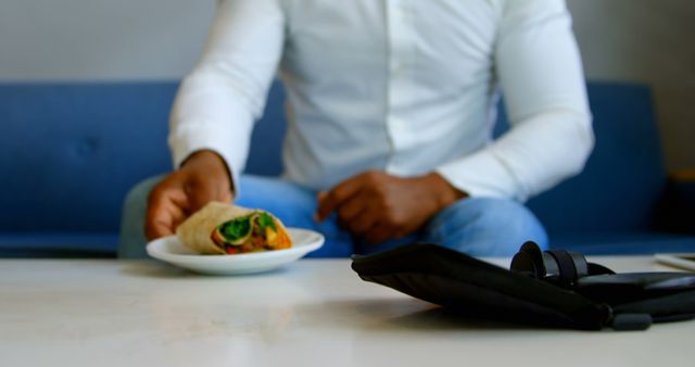 Man in white shirt and jeans reaching for a healthy wrap sandwich on a white plate. Blue sofa in background. Headphones and black case on a table in the foreground. Suitable for promoting balanced diet, casual dining, and healthy lifestyle content. Perfect for use in articles, blog posts, and advertisements related to health, wellness, and nutrition.