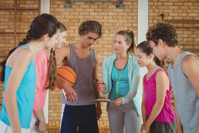 Female coach mentoring high school students in a basketball court, discussing team strategy with players holding a clipboard and basketball. Ideal for education, teamwork, youth sports, and coaching concepts. Suitable for articles, educational materials, sports training programs, and advertisements promoting youth engagement in sports.