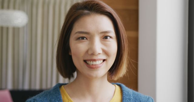 Smiling young Asian woman wearing a yellow shirt and blue sweater, standing indoors with natural light and a cheerful expression. Ideal for topics related to happiness, positivity, casual living environments, or lifestyle blog features on home decor, self-care, or modern living spaces.