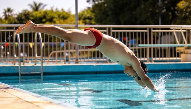Asian male swimmer diving horizontally into outdoor pool wearing red briefs, midair splash