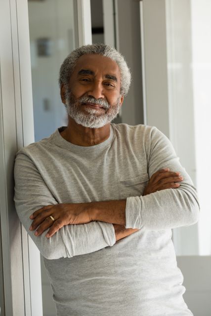 This image depicts a senior man standing with his arms crossed at the entrance of his home, smiling confidently. Ideal for use in advertisements, articles, or websites focused on senior living, retirement, health, and lifestyle. It conveys a sense of relaxation, confidence, and contentment.