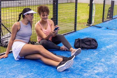 African american women relaxing on blue padel court sharing smartphone on break with duffel bag