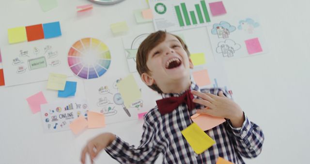 A young Caucasian boy in a plaid shirt and bow tie laughs joyfully, surrounded by colorful sticky notes and creative brainstorming materials on a whiteboard. His cheerful expression suggests a fun learning environment or a playful approach to education.