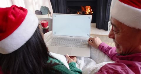 Senior couple wearing santa hats sharing laptop by fireplace, cozy holiday togetherness