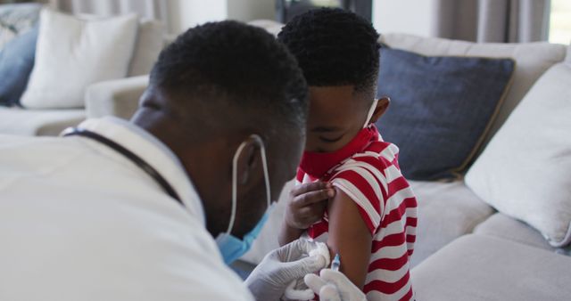 Doctor administering vaccine to young boy wearing a red striped shirt at home. The child appears calm and wears a face mask for safety. This image is ideal for illustrating healthcare services, child immunization campaigns, or promoting COVID-19 vaccination programs.