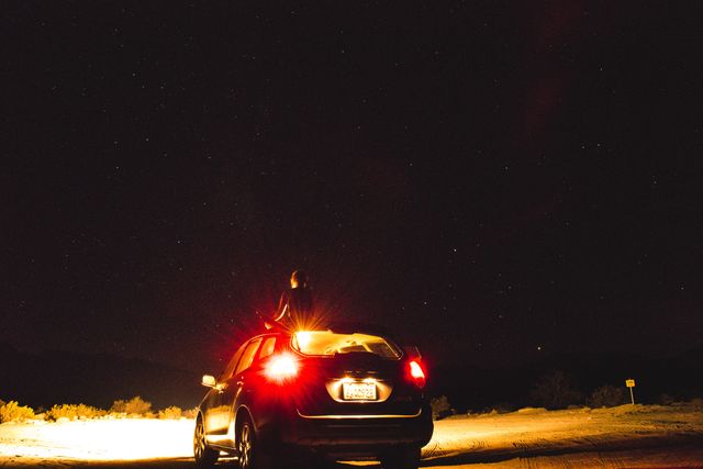 Person standing on car roof, illuminated by car headlights, gazing at night sky filled with stars. Can be used in themes of adventure, exploration, travel, solitude, and wonder.