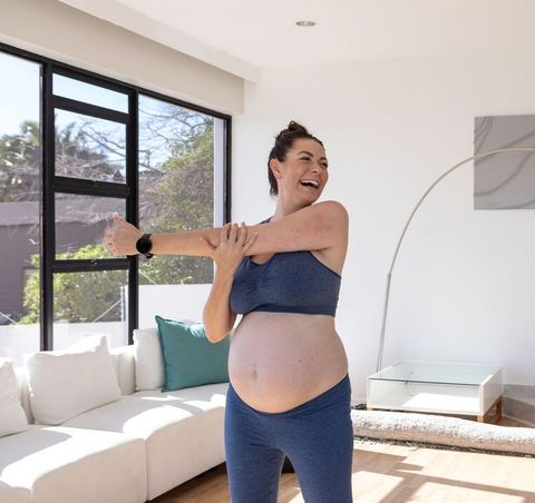 Pregnant woman stretching and laughing in sunlit modern living room wearing navy activewear