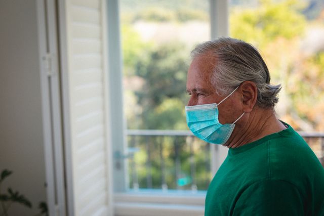 Senior man wearing a face mask standing indoors, looking out window. Useful for topics related to health, safety, elderly care, pandemic precautions, and social distancing.