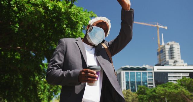 African american senior man wearing face mask holding smartphone and coffee cup hailing taxi on the road. hygiene and social distancing during coronavirus covid-19 pandemic.