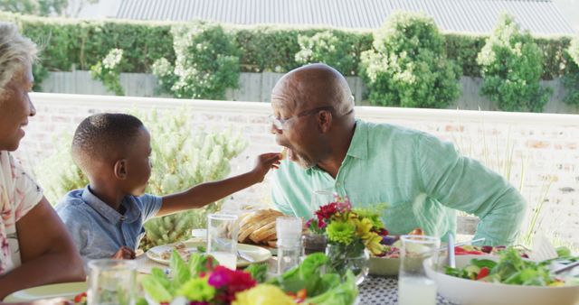 Grandfather and grandson sharing playful moment while having an outdoor meal with family during summer. Perfect for concepts related to family bonding, multi-generational interactions, outdoor dining, and fun moments.