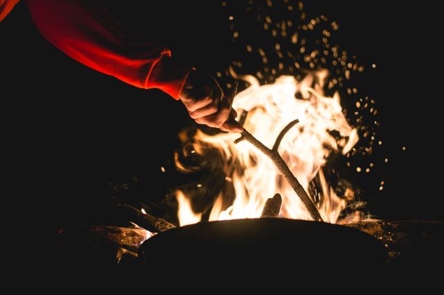 Person wearing red sleeve tending campfire with stick under night sky. Sparks illuminate darkness, creating warm, adventurous ambiance. Perfect for travel blogs, outdoor activity promotions, wilderness exploration content, or camping gear advertisements.
