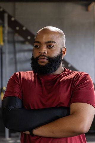 African american athlete standing arms crossed in gym wearing burgundy tee and compression sleeve