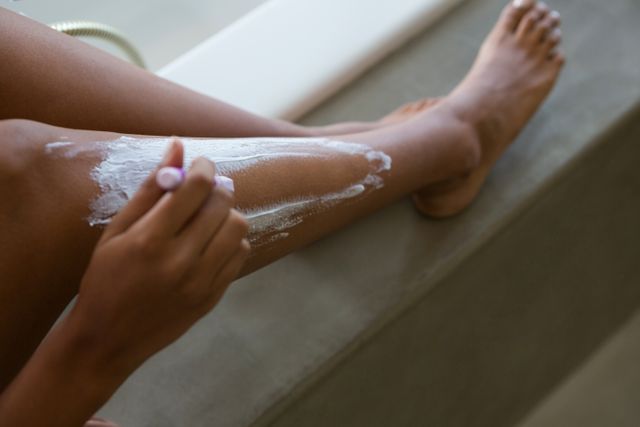 This image shows a woman shaving her leg by a bathtub, focusing on personal grooming and hygiene. It can be used for beauty and personal care advertisements, self-care articles, or tutorials on shaving techniques.