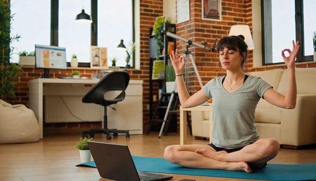 Woman meditating on yoga mat in home workspace with laptop practicing mindfulness