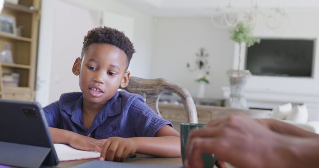 A young African American boy in a blue shirt sits at a table, engrossed in studying with a digital tablet. He is possibly participating in an online class or doing homework. This depicts modern education, use of technology in learning, and home study environment. Ideal for illustrating online education platforms, homeschooling, educational technology uses, and young students’ learning process at home.