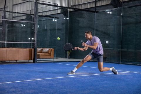 Male athlete lunging on blue padel court hitting ball with black racket indoor action