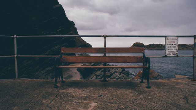 Bench located along a coastal path, offering a tranquil view of the ocean. Gray sky and wooden bench emphasize solitude and calmness. Ideal for themes related to relaxation, contemplation, solitude, and natural beauty.