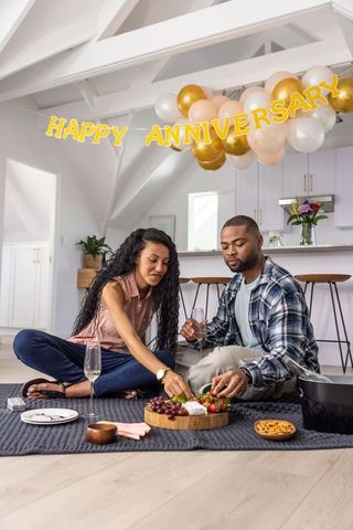 Black couple sharing charcuterie and wine on rug under happy anniversary balloons