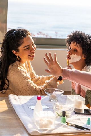 Diverse female friends applying creamy face mask with brush by sunlit ocean-view window