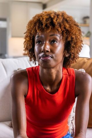 Contemplative african american woman sitting on sofa in red sleeveless top gazing up calmly