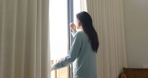 Asian woman standing at sliding glass door shielding eyes with curtains morning light