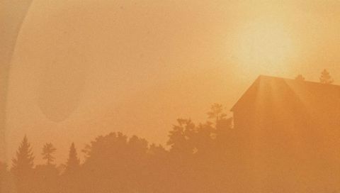 Glowing sun casting golden rays over silhouetted house and conifer treeline at hazy dusk