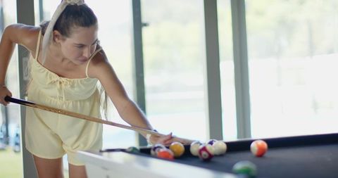 Young woman aiming cue while playing pool in sunlit modern interior with copy space