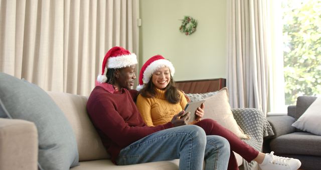 Couple wearing Santa hats sitting on couch with digital tablet. Suitable for holiday season promotional content, digital Christmas cards, online family celebrations, and festive marketing materials.