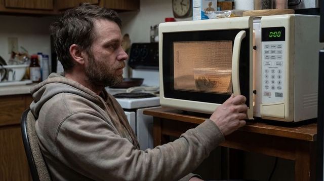 Tired bearded man sitting at kitchen chair warming food in microwave in cluttered home