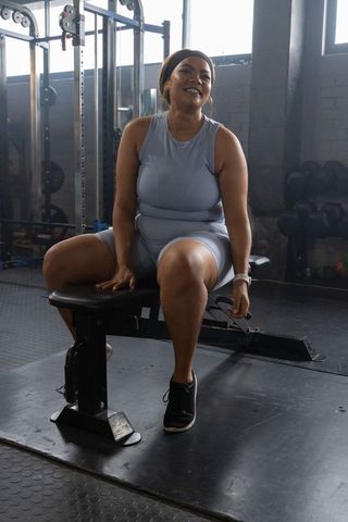 African american woman sitting on bench in gym wearing light blue athletic set smiling
