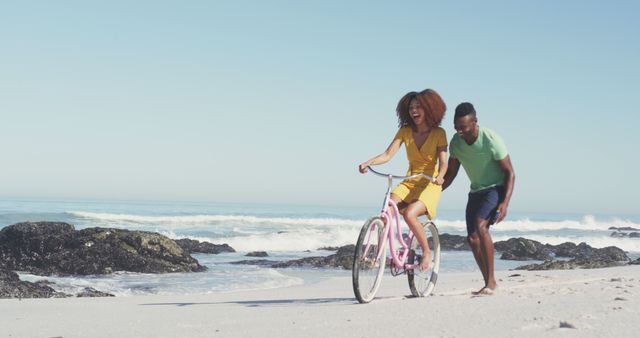 A cheerful couple is enjoying a sunny day on the beach. The woman is riding a bicycle on the sand while the man jogs alongside her, both smiling and laughing. The ocean waves provide a scenic background, creating a perfect summer atmosphere. This image could be used in advertisements or promotions related to travel, vacation, leisure activities, health and fitness, and outdoor recreation.