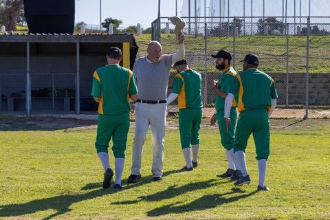 Coach raising cap acknowledging baseball team in green uniforms on sunlit field