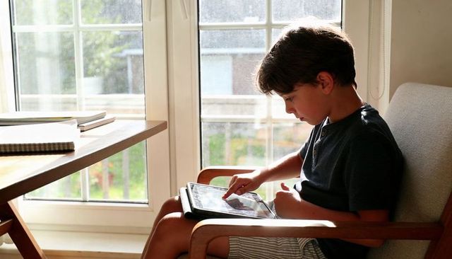 Boy using tablet in sunlit armchair by window, focused digital learning and relaxed play