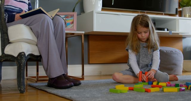 Girl playing with building blocks in living room at home. Grandmother reading a book while granddaughter playing.