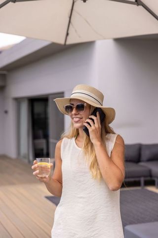 Smiling woman wearing straw hat talking on phone holding lemon drink on sunlit deck