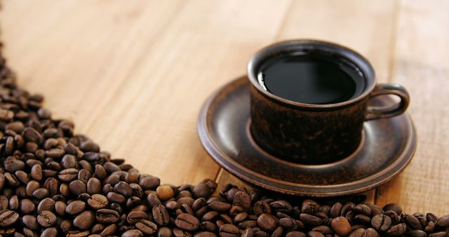 Rustic coffee cup filled with dark coffee surrounded by scattered coffee beans on wooden table. Perfect for use in coffee shop promotions, breakfast advertisements, food and drink blogs, and product packaging.