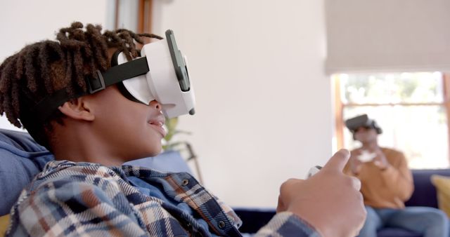 Young African American boy engaged in a VR game using a headset and controller while sitting on a cozy sofa at home. Perfect for illustrating modern gaming, technology use, child entertainment, and family activities. Suitable for advertisements, blog posts, and articles about gaming technology and virtual reality.