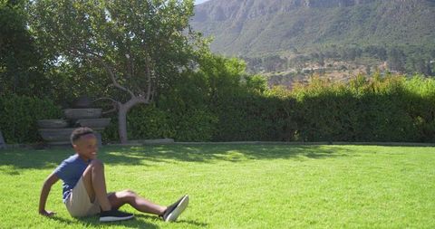 African boy relaxing on lawn in sunny backyard with mountain view, wearing blue tee and sneakers