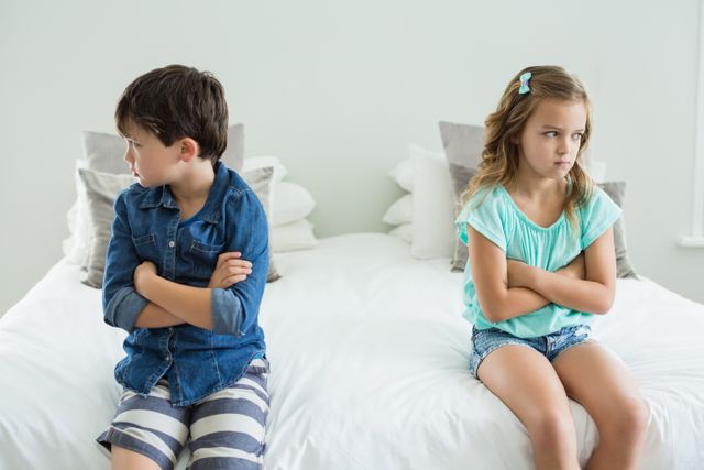 Two children, a boy and a girl, are sitting on a bed with their arms crossed, looking upset. They are in a bedroom at home, indicating a moment of conflict or disagreement. This image can be used to illustrate themes of sibling rivalry, family dynamics, childhood emotions, or conflict resolution in educational materials, parenting blogs, or family counseling resources.