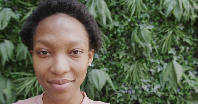 Portrait of an African woman with short hair, smiling outdoors by green plants. Perfect for use in projects related to nature, wellness, outdoor activities, environmental consciousness, and natural lifestyle imagery.