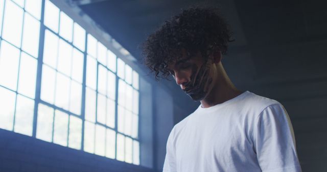 This image shows a young man with curly hair, wearing a white shirt, in an industrial setting with large windows. His thoughtful and contemplative expression adds an edgy mood to the scene. Ideal for use in campaigns related to thinking, introspection, modern industry, or contemporary fashion.