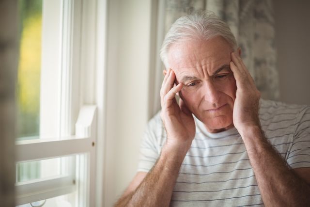 Elderly man standing by window with head in hands, appearing stressed and worried. Useful for topics related to mental health, aging, loneliness, and emotional well-being. Can be used in articles, blogs, and advertisements focusing on senior care, mental health awareness, and support for the elderly.