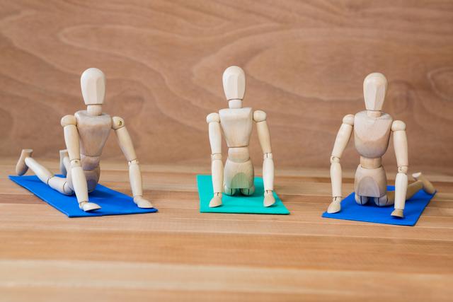 Wooden mannequins are shown practicing yoga poses on colorful mats against a wooden background. This image can be used for promoting yoga classes, fitness routines, wellness programs, or illustrating concepts of flexibility and health.