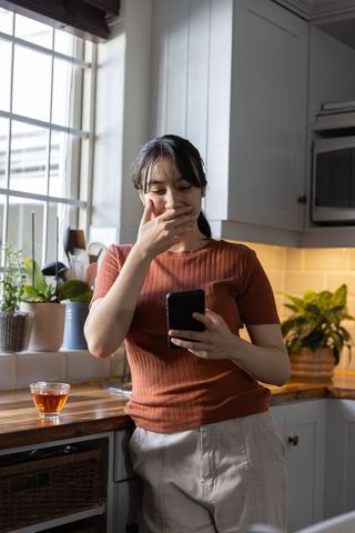 Surprised asian woman reading smartphone in bright kitchen covering mouth, candid lifestyle