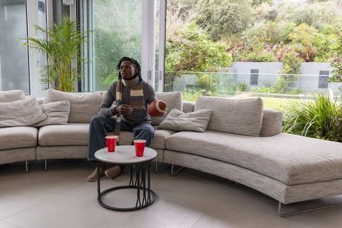African american man relaxing on gray sofa holding football and checking phone