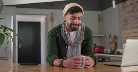 Indian man leaning on wooden kitchen island holding mug near laptop, cozy remote work
