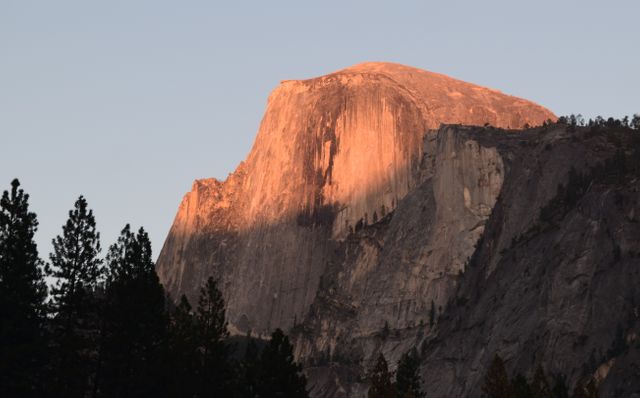 The golden light of the sunset illuminates the iconic Half Dome in Yosemite National Park. Silhouettes of evergreen trees frame the majestic, granite formation. This image is ideal for promoting national parks, appealing to hikers and nature enthusiasts, and illustrating travel brochures. It captures the serene and timeless beauty of one of the most famous landmarks in America's natural landscape.