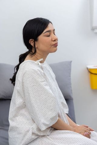 Calm woman sitting on exam bed wearing hospital gown during routine clinic checkup